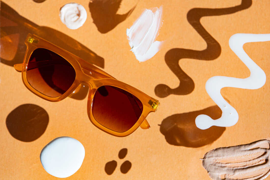Photograph of various sunscreens squirted out on a piece of glass that's elevated above an orange background, creating shadows. There is a pair of sunglasses arranged between the sunscreen dollops.