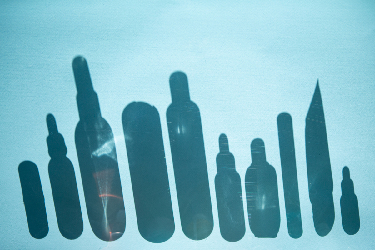 Photograph the shadows of ten skincare product bottles of varying sizes and shapes on an aqua colored backdrop.
